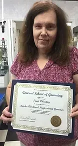Woman holding a grooming certificate from Concord School of Grooming, smiling, in a shop with checkered floor.
