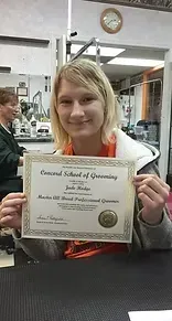 Woman holding certificate from Concord School of Grooming, smiling in a salon.
