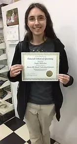 Woman holding a certificate, smiling, in a shop, wearing glasses, black coat, beige pants.
