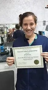 Woman holds a certificate from the Concord School of Grooming. She smiles in a grooming shop setting.