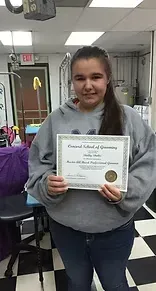 Woman holding a certificate from a school of grooming, smiling. She's in a grooming shop wearing a gray sweatshirt and jeans.