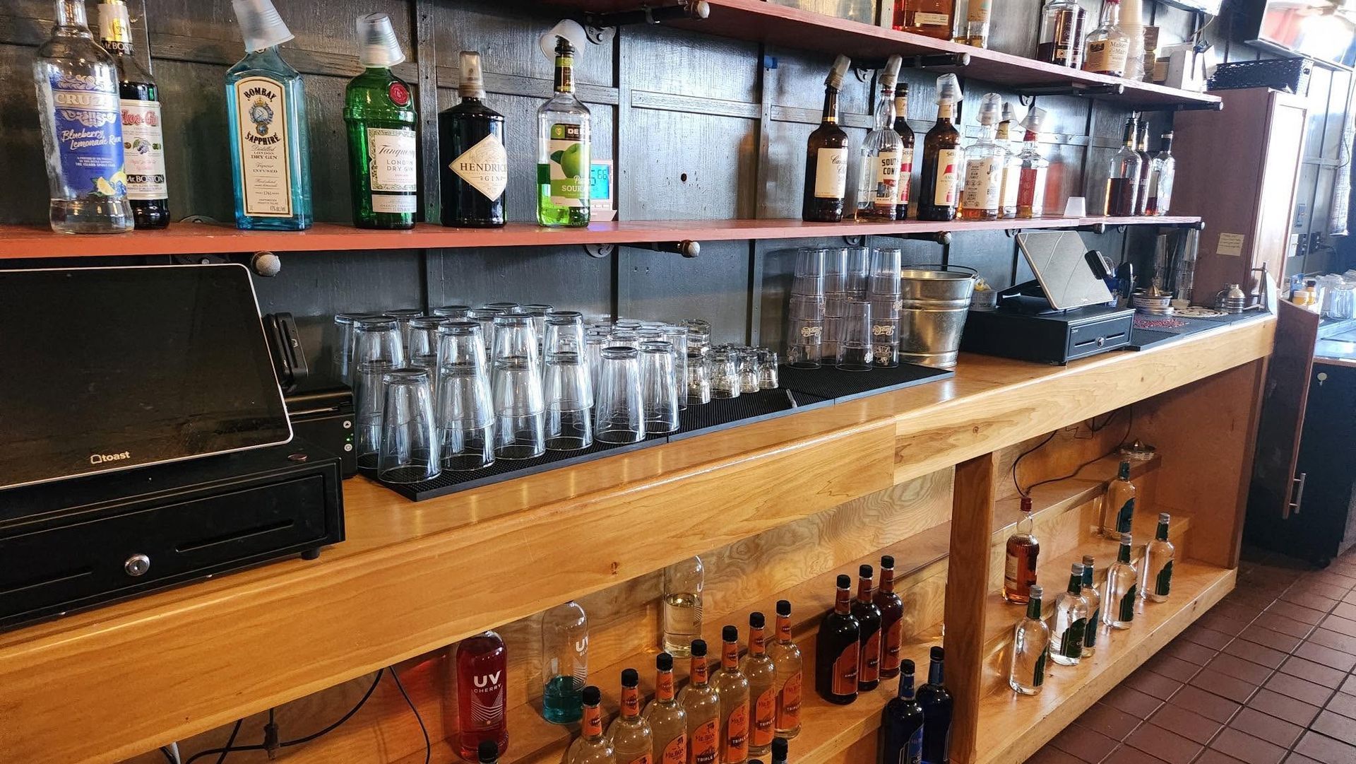 Bar with liquor bottles, glassware, and a cash register on a wooden counter, viewed from the side