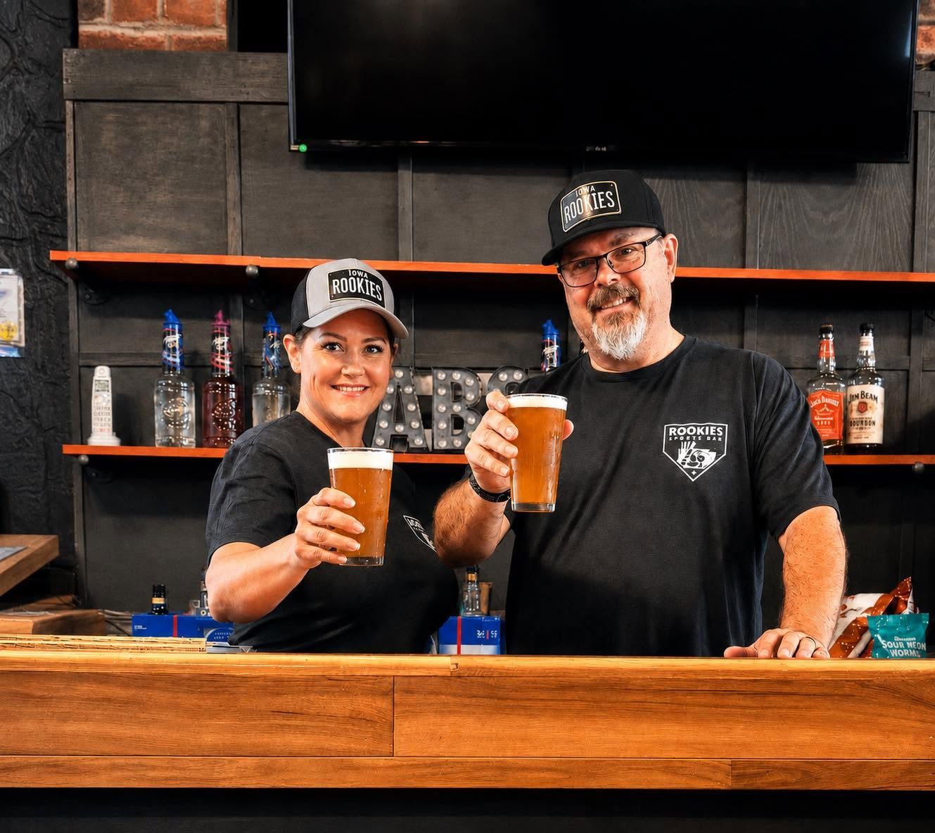 Two people in black shirts toast with beer glasses behind a wooden bar.
