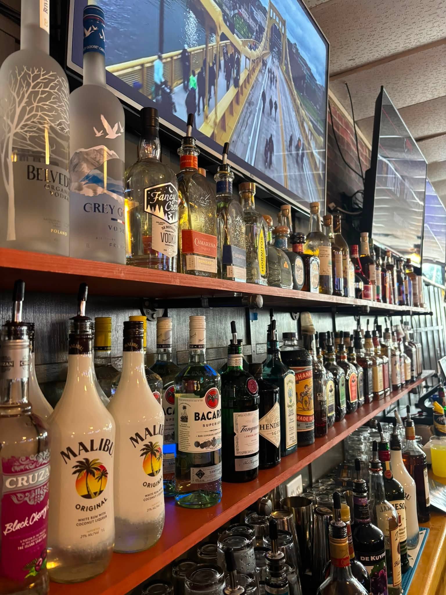 Rows of liquor bottles on a bar shelf with a TV above showing a bridge scene.