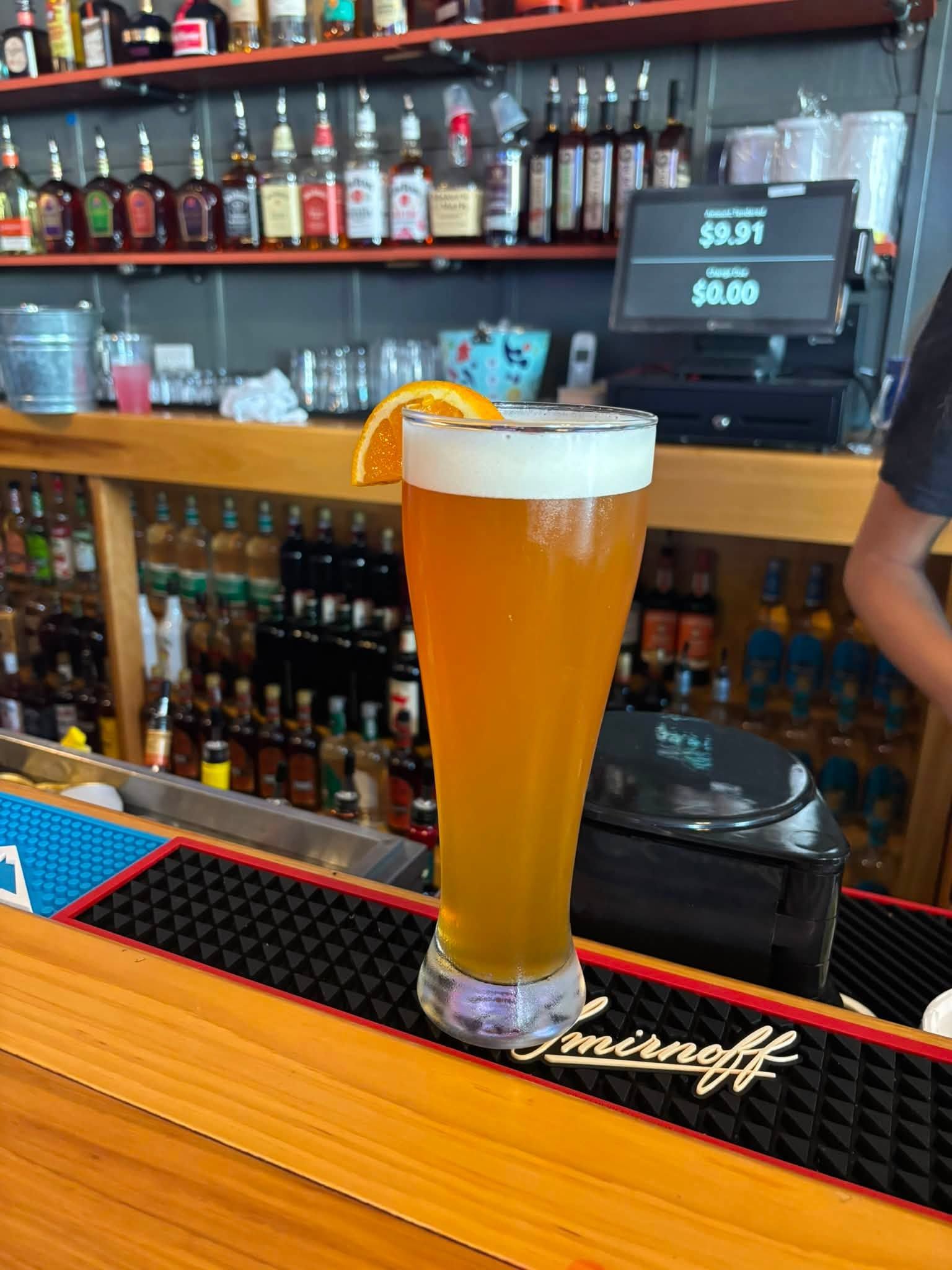 Tall orange beer with a salted rim and orange garnish on a bar counter, with bottles and taps behind.