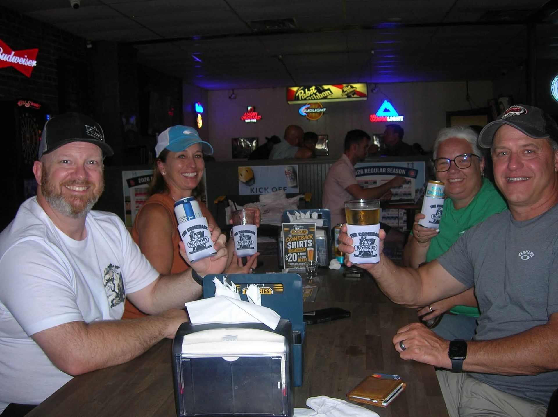Five friends at a bar table raising branded mugs, smiling under neon lights