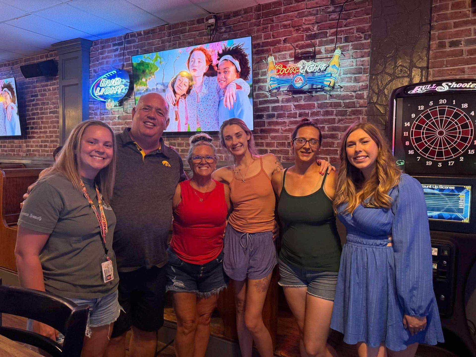 Group posing together in a brick-walled bar, smiling in front of a TV and dartboard.