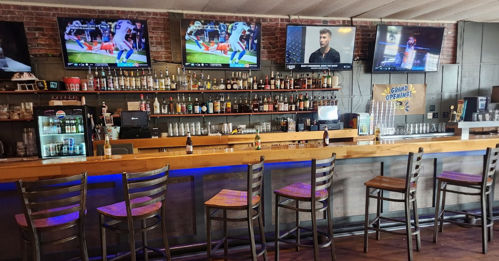 Empty sports bar with wooden counter, high stools, shelves of bottles, and TVs showing games.