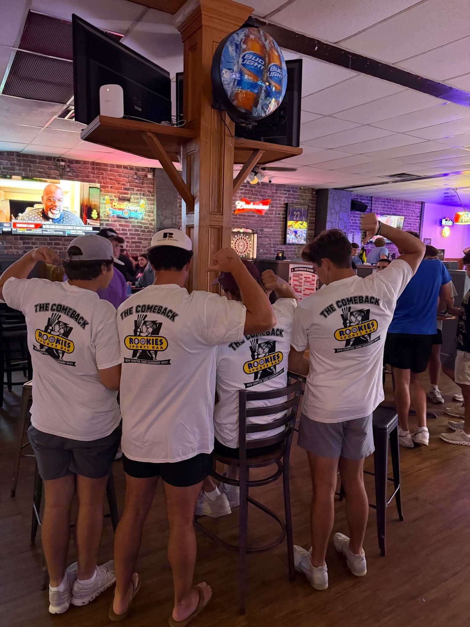 People in white shirts gathered at a bar, watching a TV under a circular sign in a lively indoor venue.