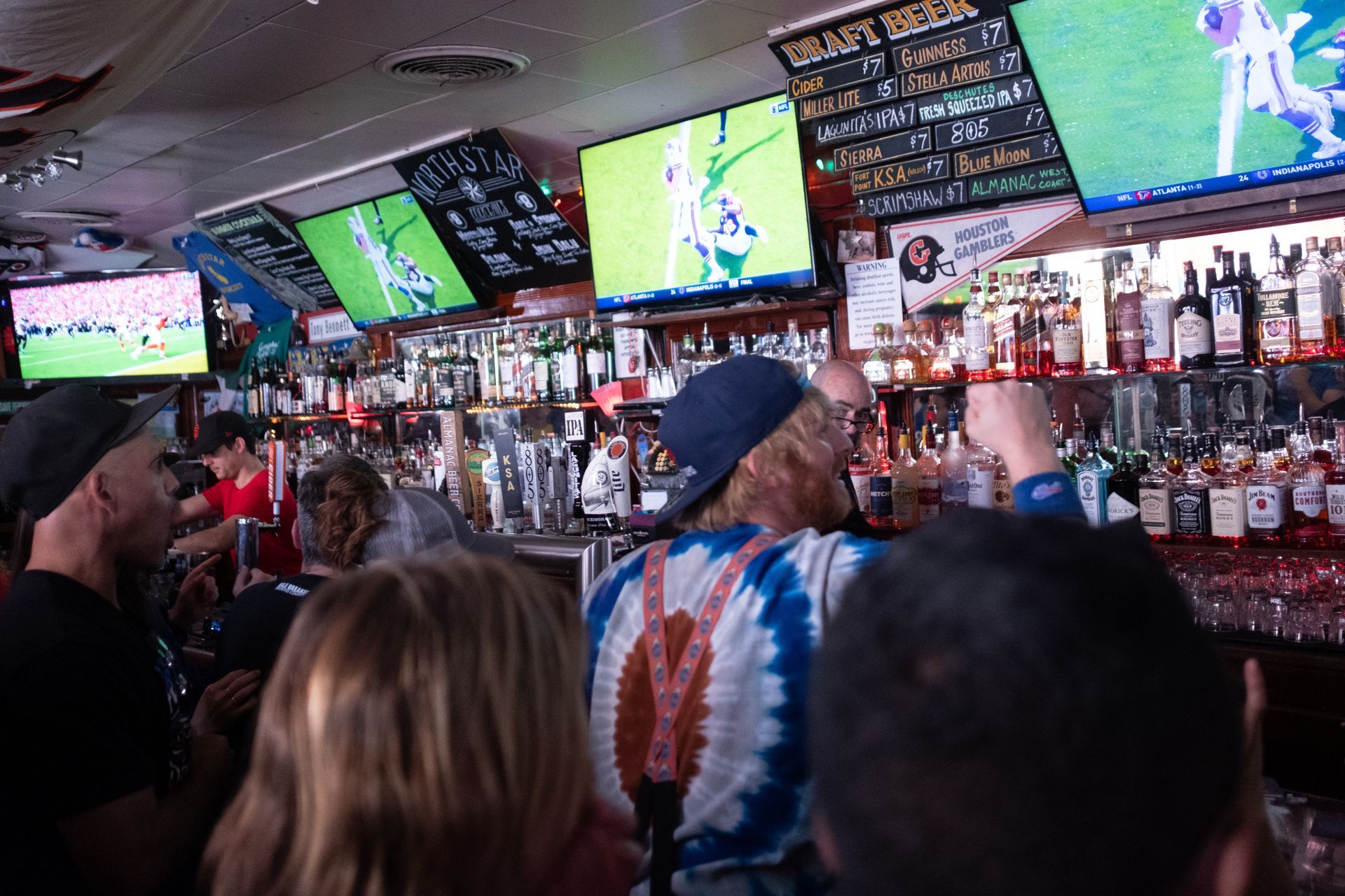 Crowded sports bar with patrons watching multiple TVs showing a game, bar shelves in the background