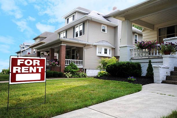For rent sign in front of a beige suburban house with a driveway and porch
