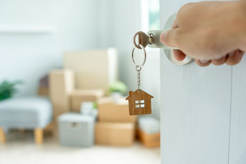 Hand holding a key with a small house keychain in front of moving boxes in a bright room