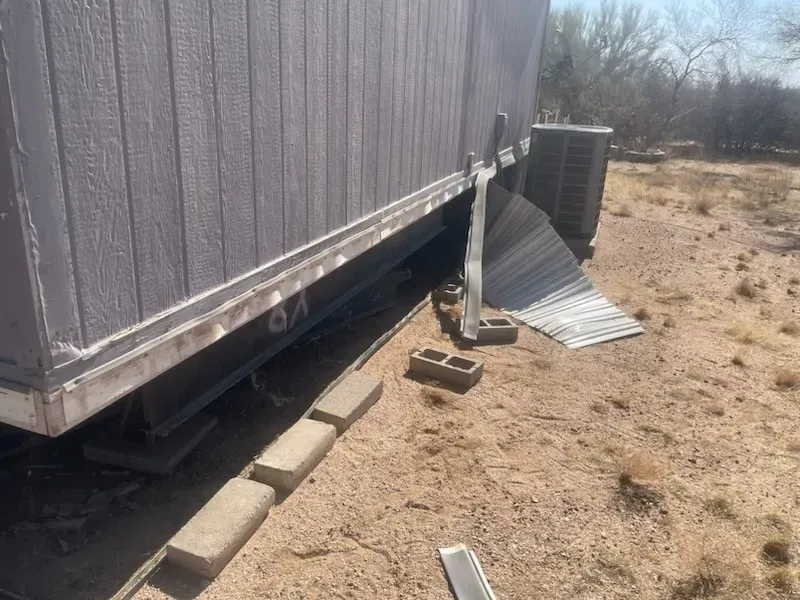 Gray-sided mobile home with detached metal paneling and air conditioning unit on a dirt lot.