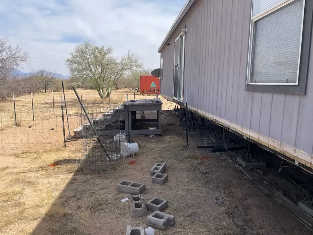 Side view of a house with visible underpinnings; cinder blocks and steps are present in a dirt yard.