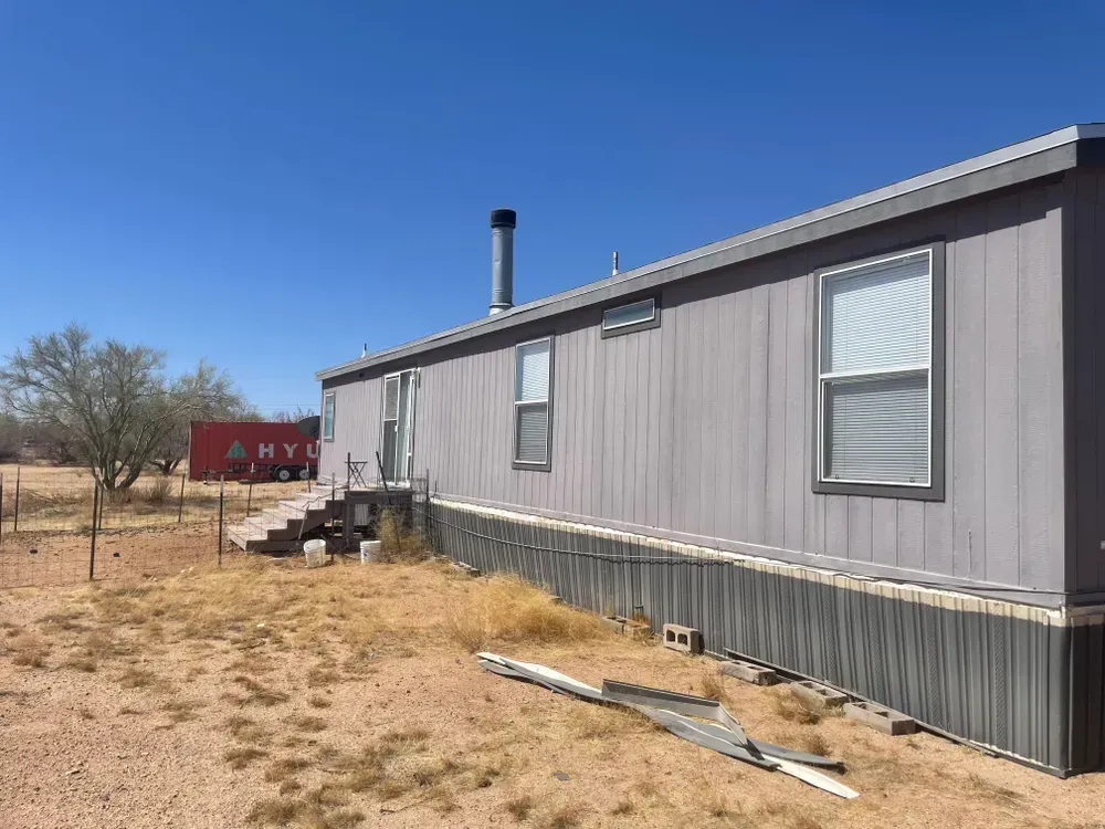 Gray mobile home with stairs, chimney, and windows in a dry, outdoor setting. Red building in the distance.