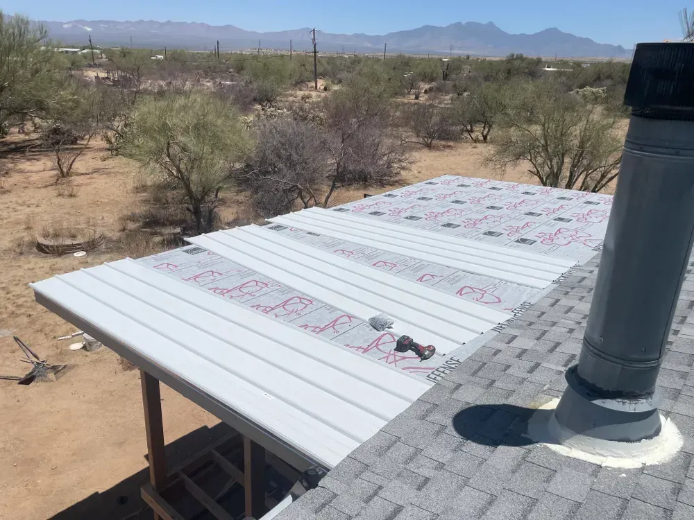 A partial roof with metal panels being installed, adjacent to an existing shingled roof, in a desert setting.