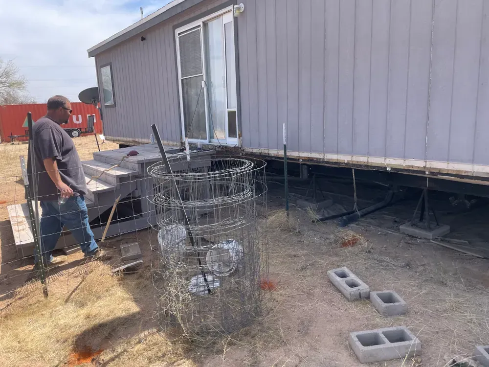 Man near house with wire cage, cinder blocks, and steps; sunny outdoors.