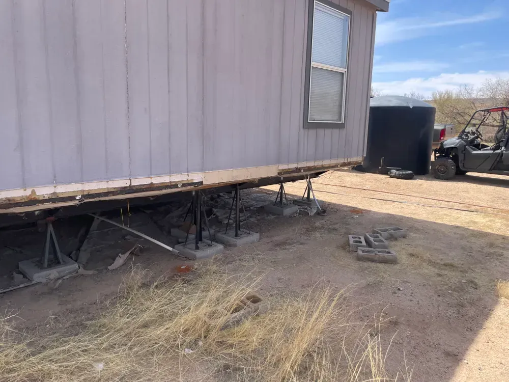 Mobile home elevated on concrete blocks and metal supports. Outside setting with dry brush and vehicles.