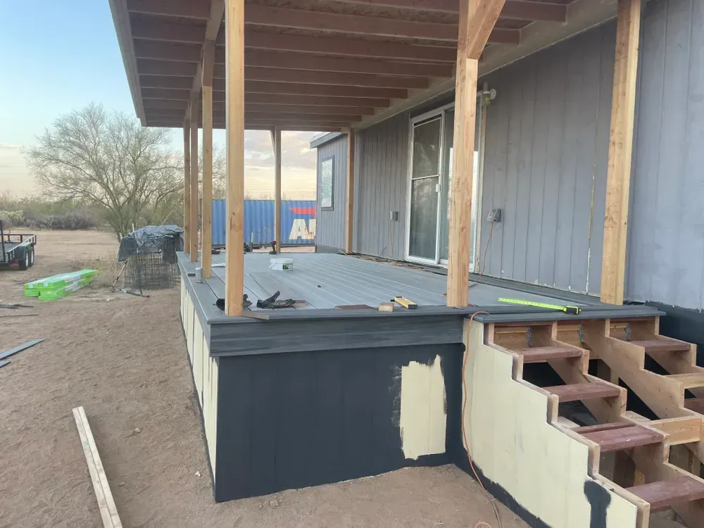 A house with a partially constructed deck and stairs. Wooden supports frame the roof, gray siding, desert setting.