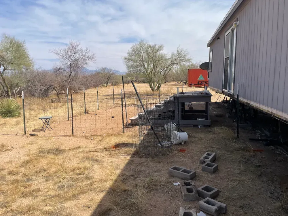 Backyard with a light-colored mobile home, chicken coop, fence, and sparse vegetation under a partly cloudy sky.