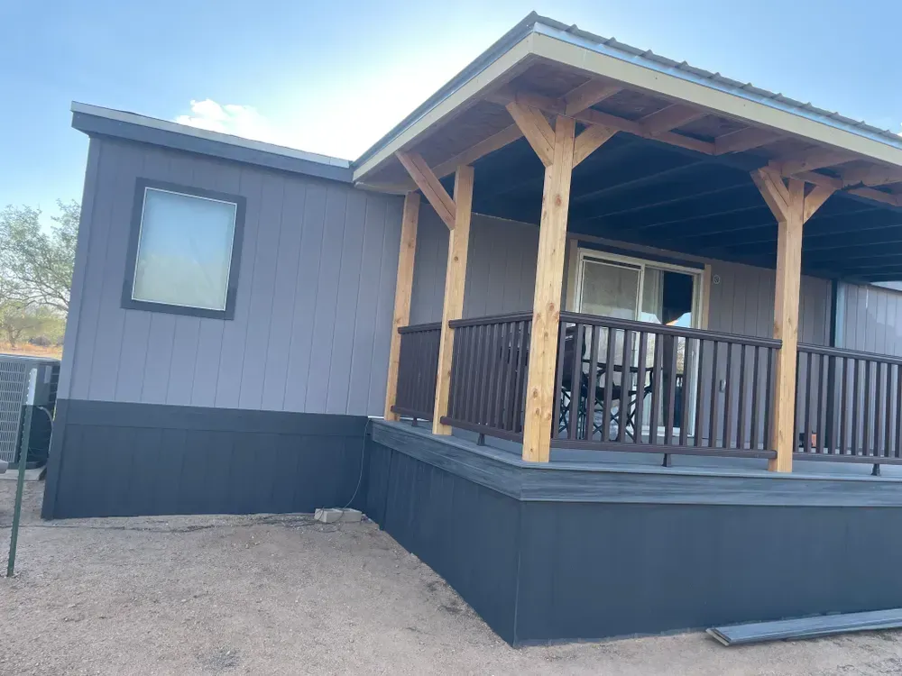 Gray and dark gray house with a wooden porch and railing, set on gravel.
