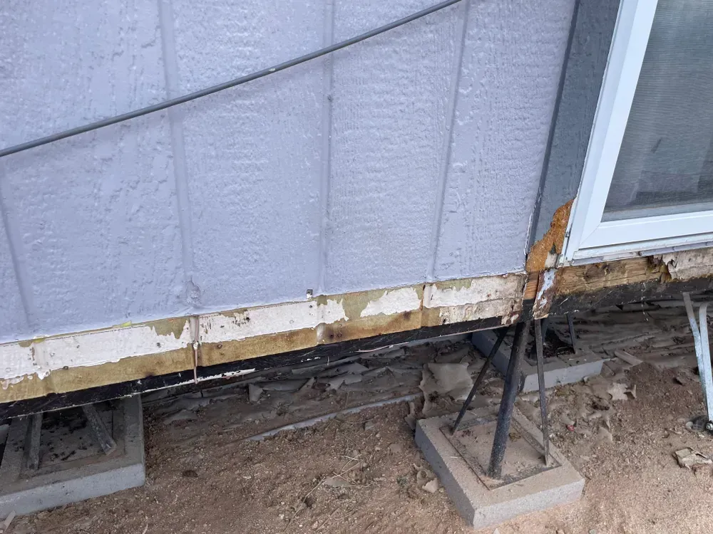 Damaged house exterior with peeling paint, exposed wood, and concrete support blocks.