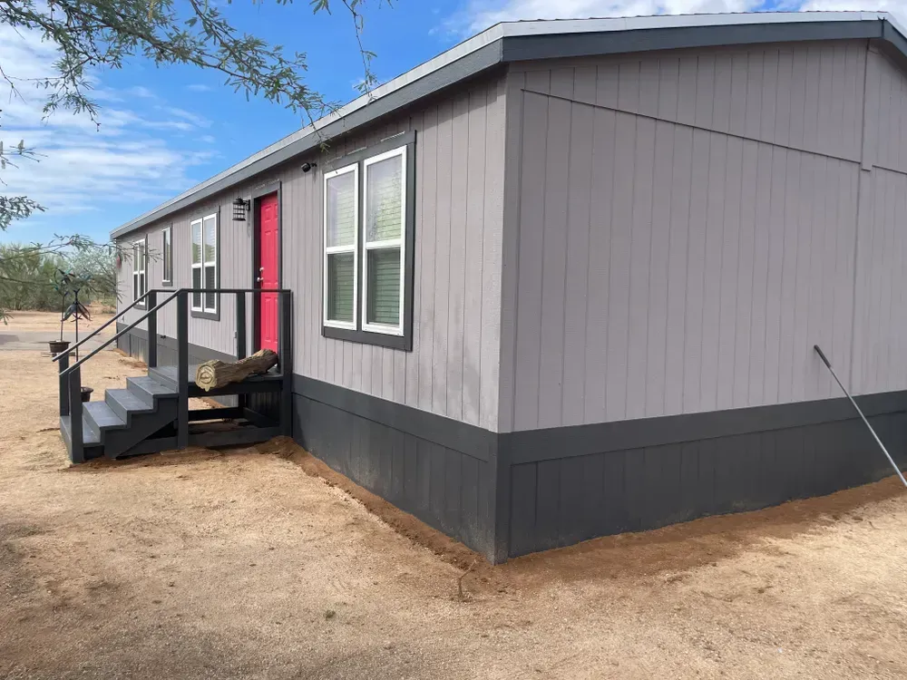Gray and black mobile home with red door, steps leading to entrance. Sunny day, desert setting.