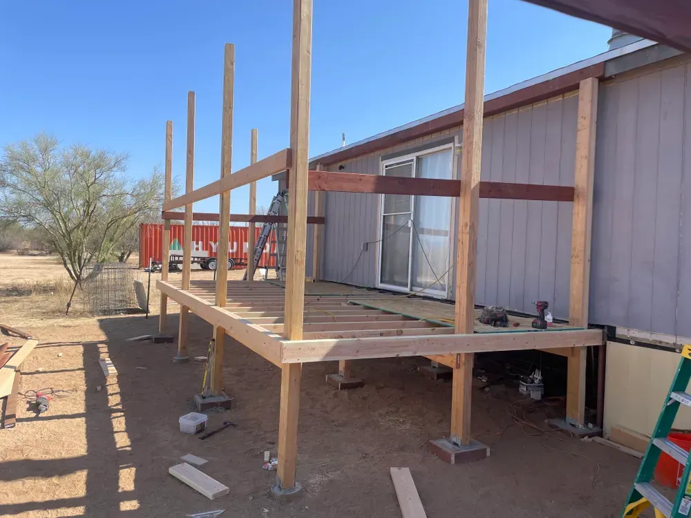 Deck construction in progress; wooden frame built against a light gray building with a sliding door.