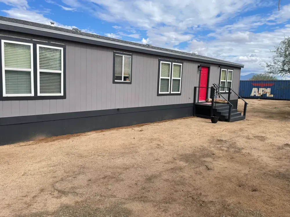 Gray mobile home with red door, black trim, and white-framed windows on a sandy lot; blue sky.