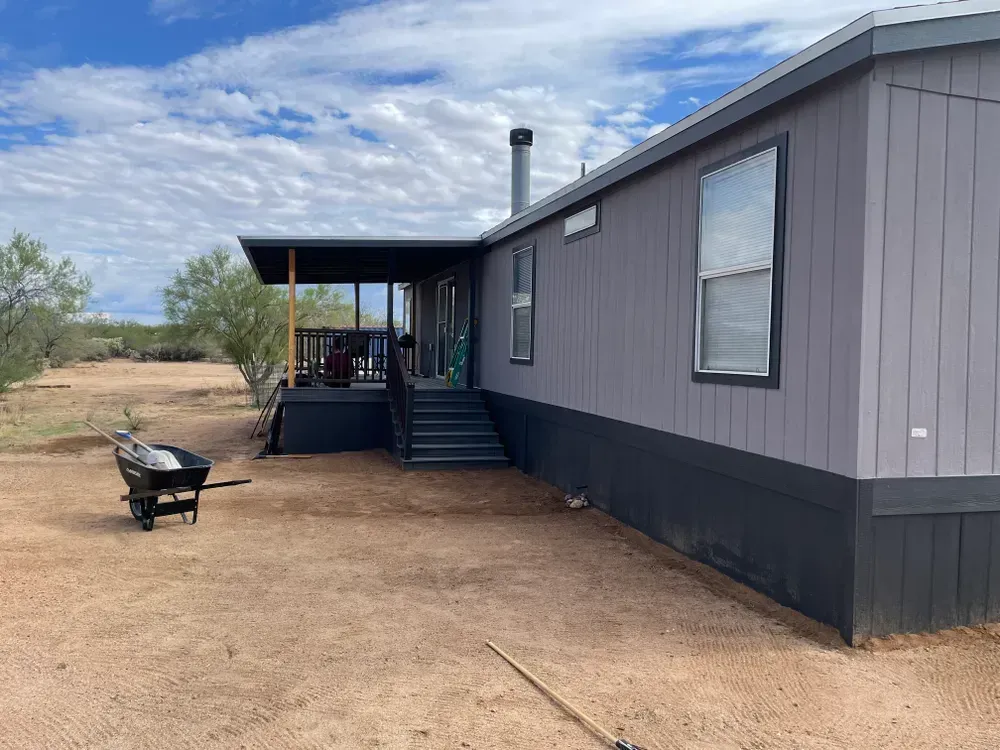 Gray mobile home with porch and steps, set on a dirt lot under a cloudy blue sky.