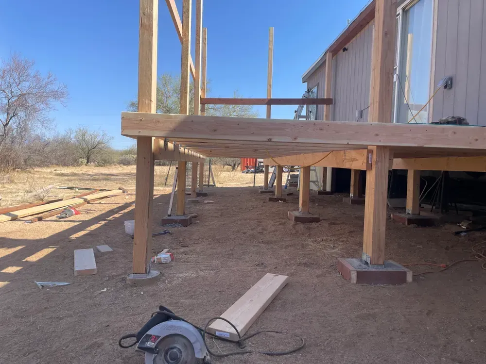 Deck under construction, wooden beams and supports in a yard next to a house. Saw and lumber in the foreground.