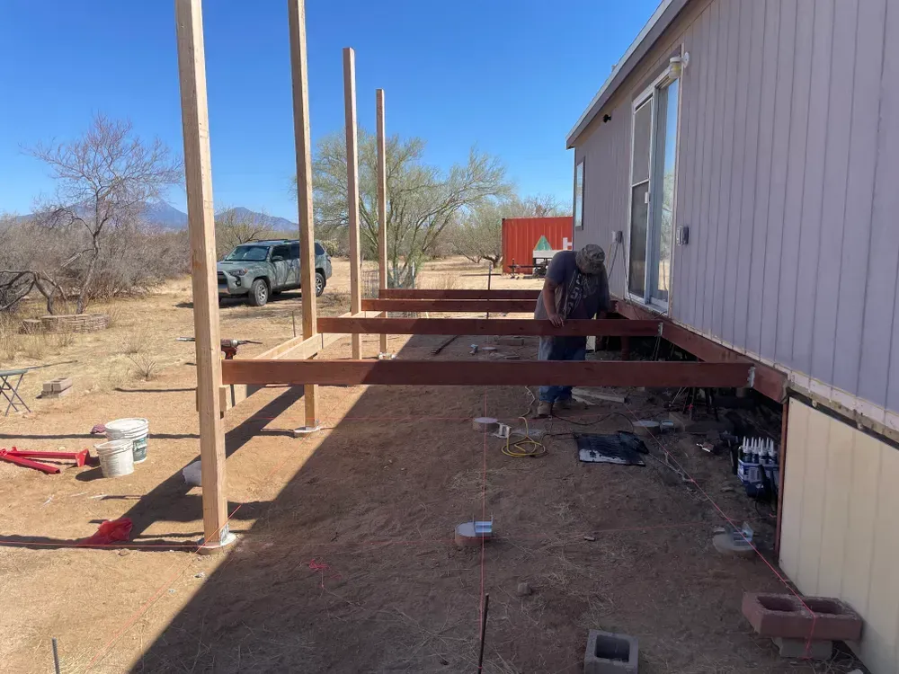 Construction worker building a deck next to a house in an outdoor, desert-like setting.