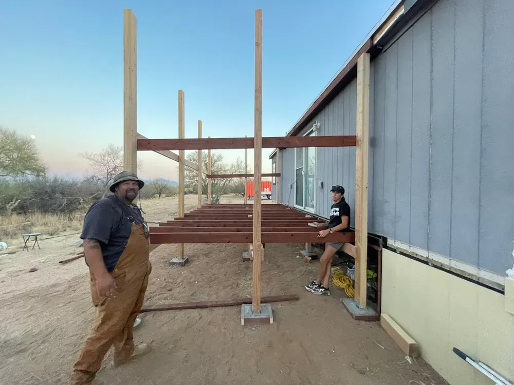 Two people constructing a wooden deck attached to a gray house; desert landscape in background.