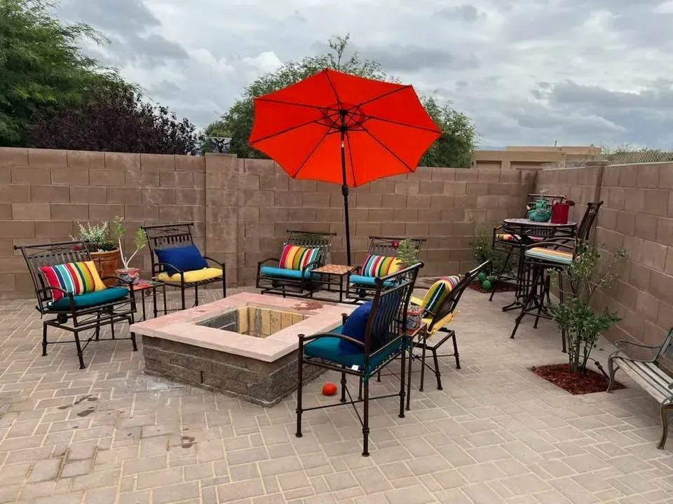 Outdoor patio with fire pit, seating, and orange umbrella against a block wall under a cloudy sky.