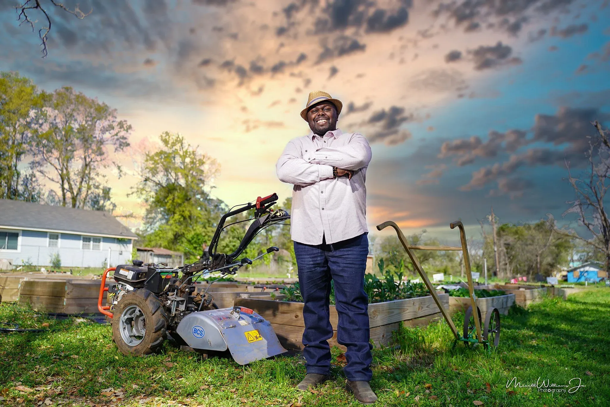 Farmer Fredo smiling with arms crossed beside a tiller and raised garden beds at sunset