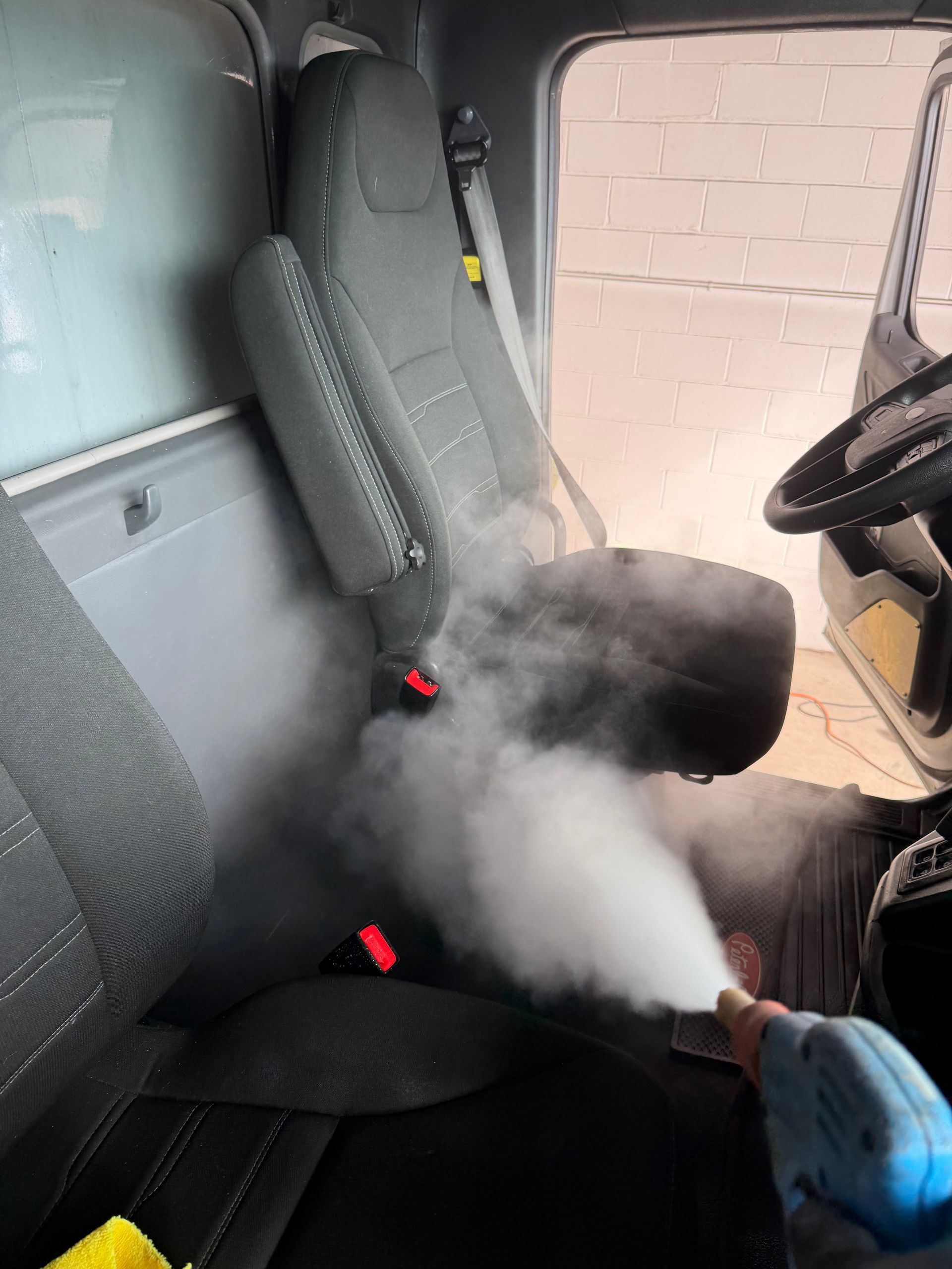 Steam cleaning a truck cab seat with a handheld tool, white vapor filling the gray interior.