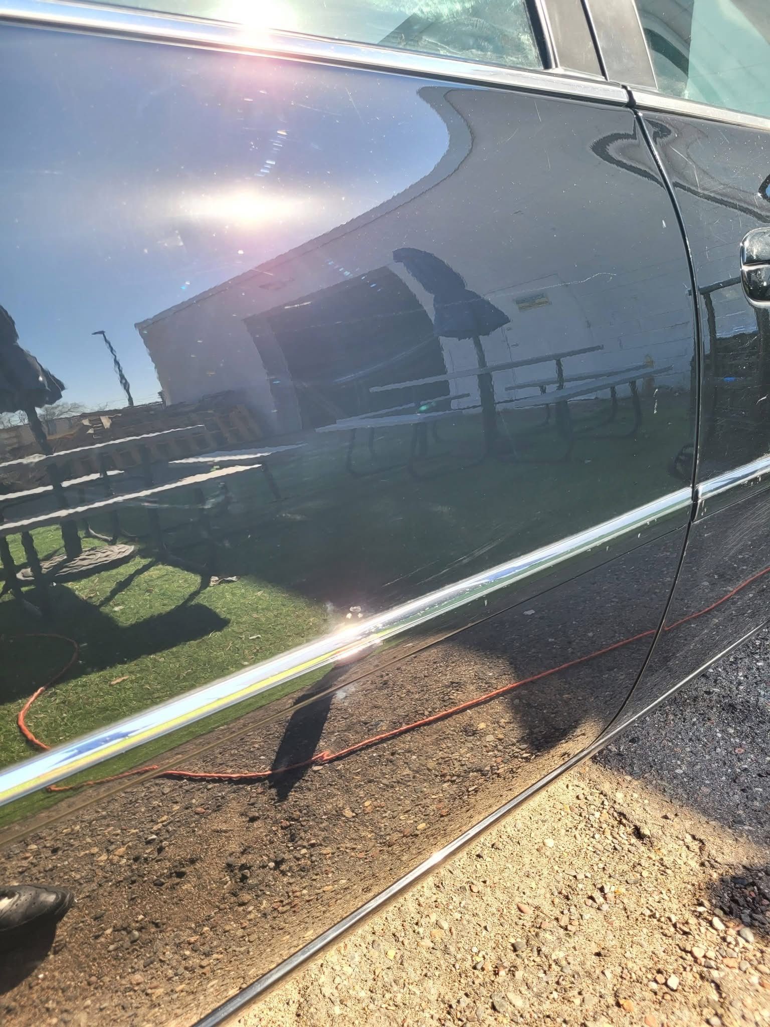 Close-up of a dark car door with reflections of a house and fence in bright sunlight.