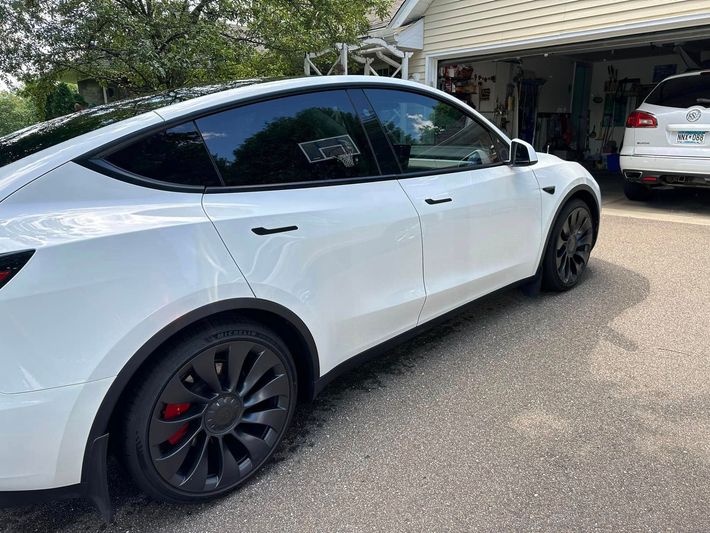 White Tesla SUV parked in a driveway beside a house, rear side view.
