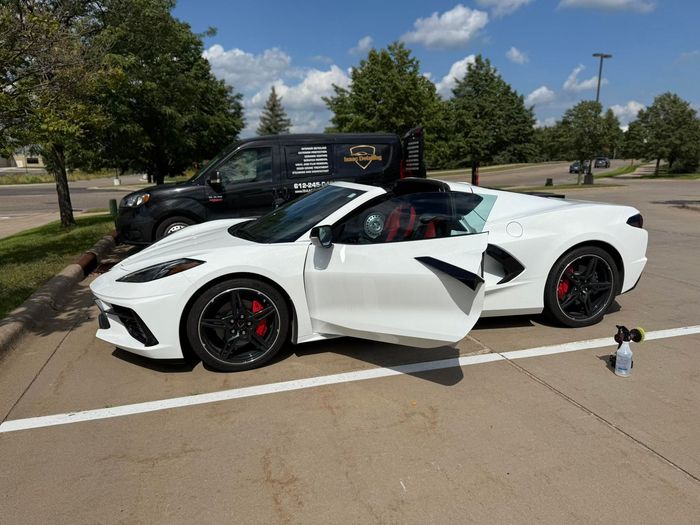 White sports car with open door parked in a lot beside a black SUV under trees.