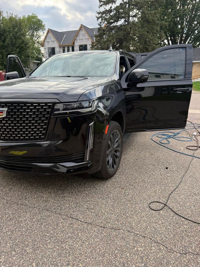 Black SUV parked on a driveway with the driver door open and a hose on the ground near a house.