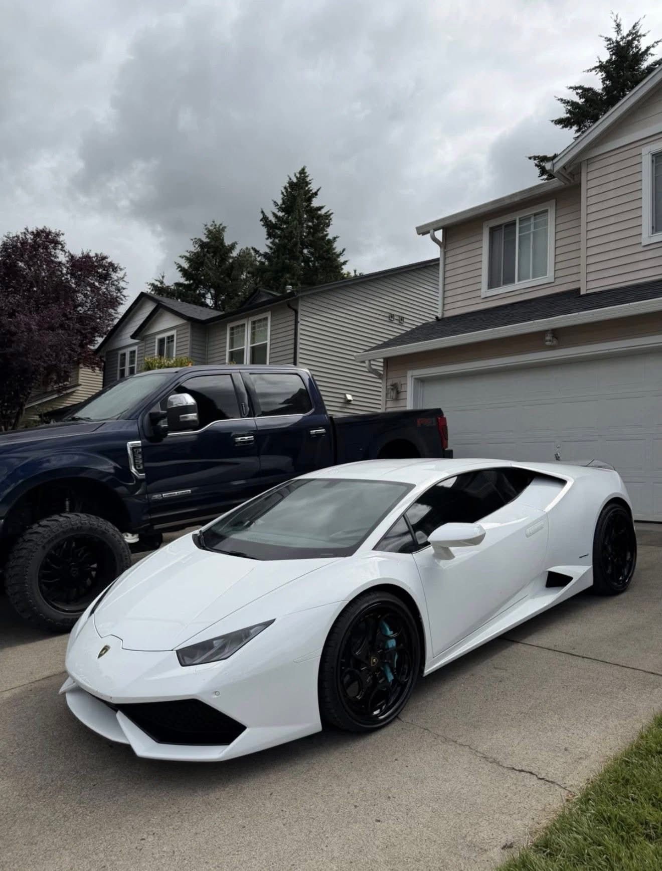 White Lamborghini parked in a suburban driveway beside a lifted black pickup truck, under cloudy skies