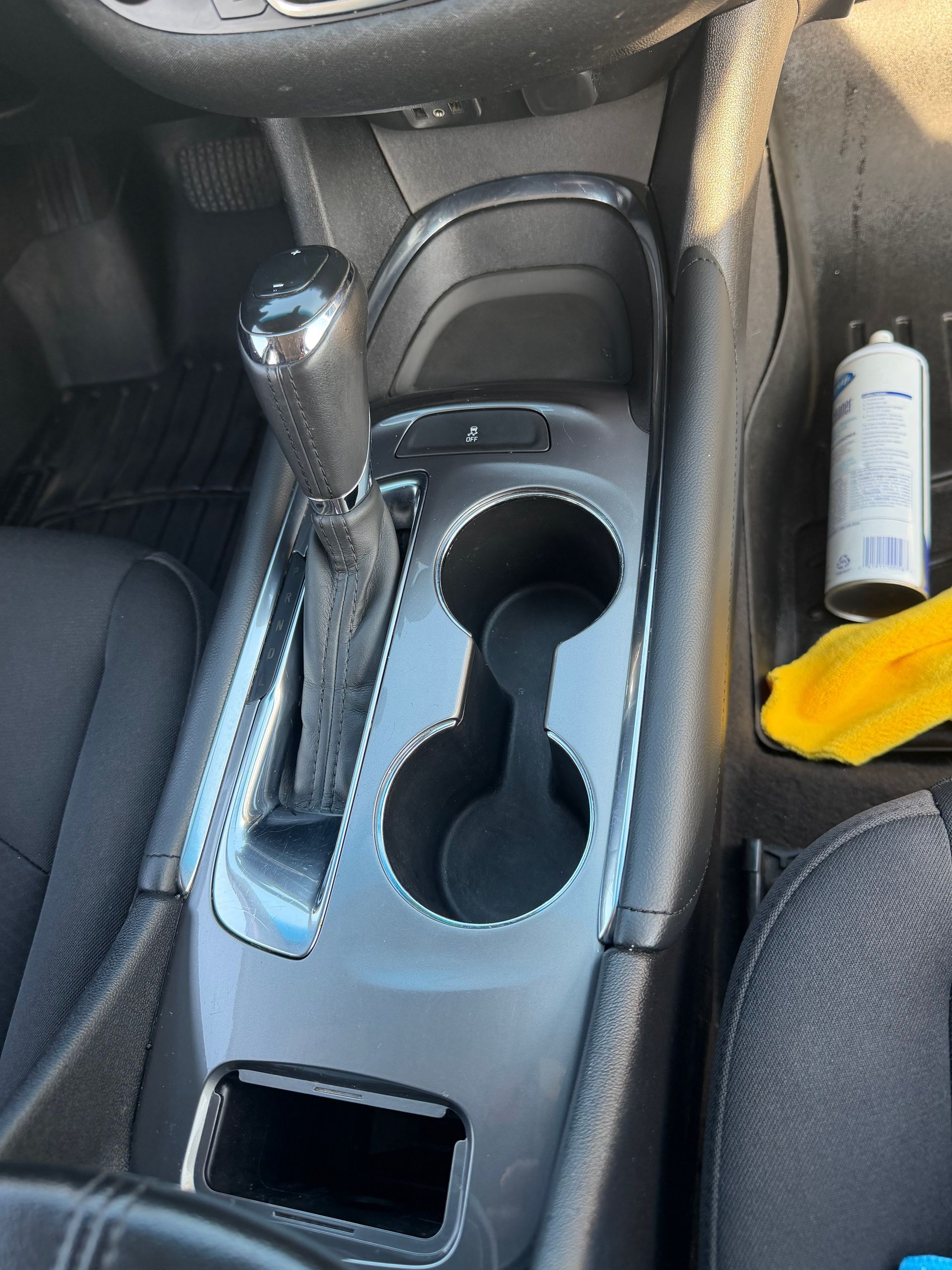 Car center console with shifter, cupholders, and a small rectangular storage opening; yellow cloth on passenger side.