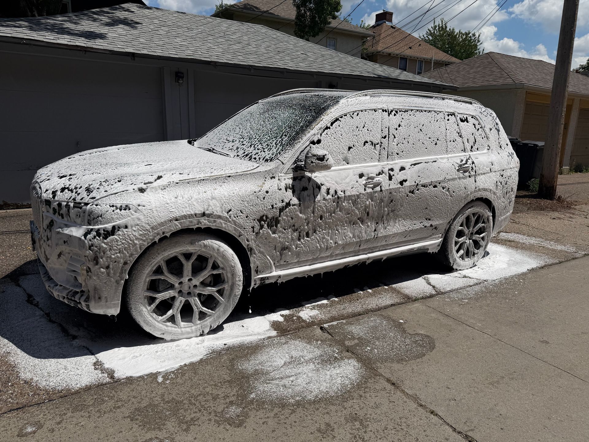 White SUV covered in foam, parked in a driveway beside a garage during a car wash.