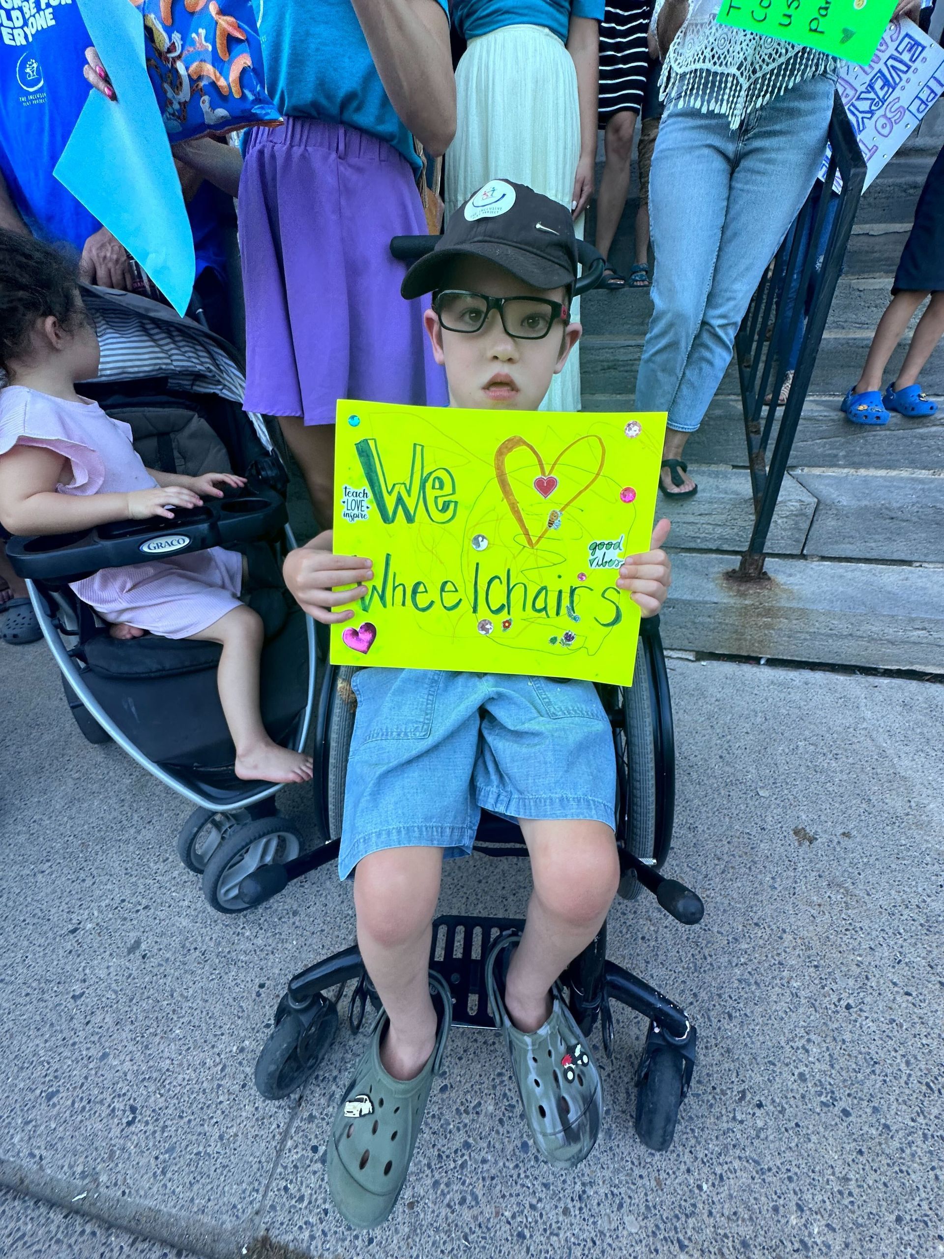 A young boy is sitting in a wheelchair holding a sign.