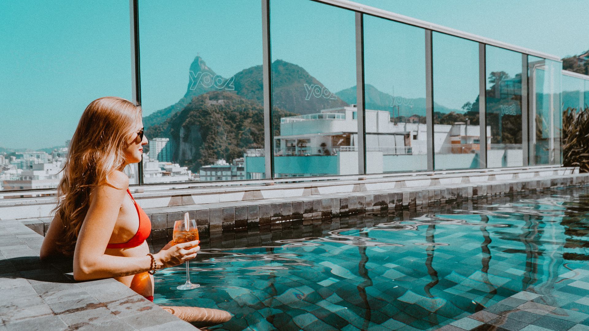 Mulher de biquíni vermelho toma drinque em uma piscina na cobertura, com vista para as montanhas do Rio de Janeiro.