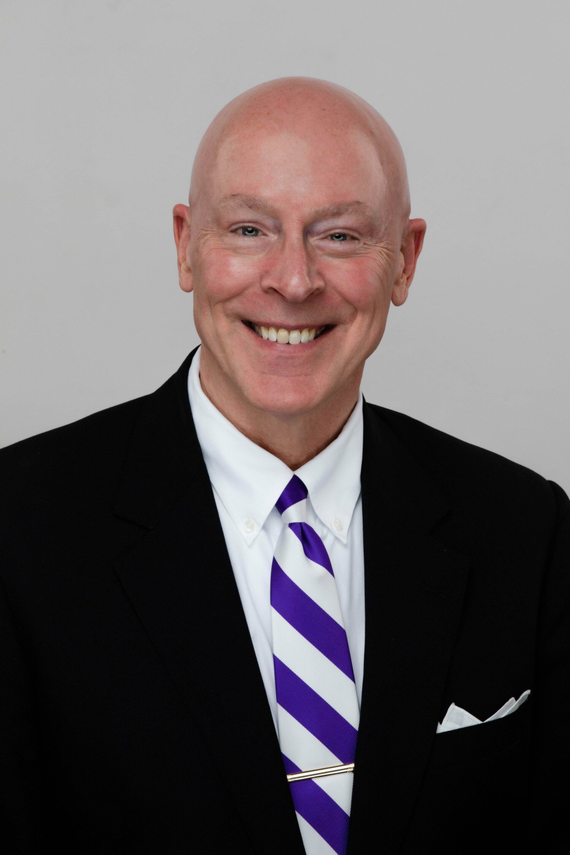 Smiling bald man in a black suit with a purple striped tie on a gray background