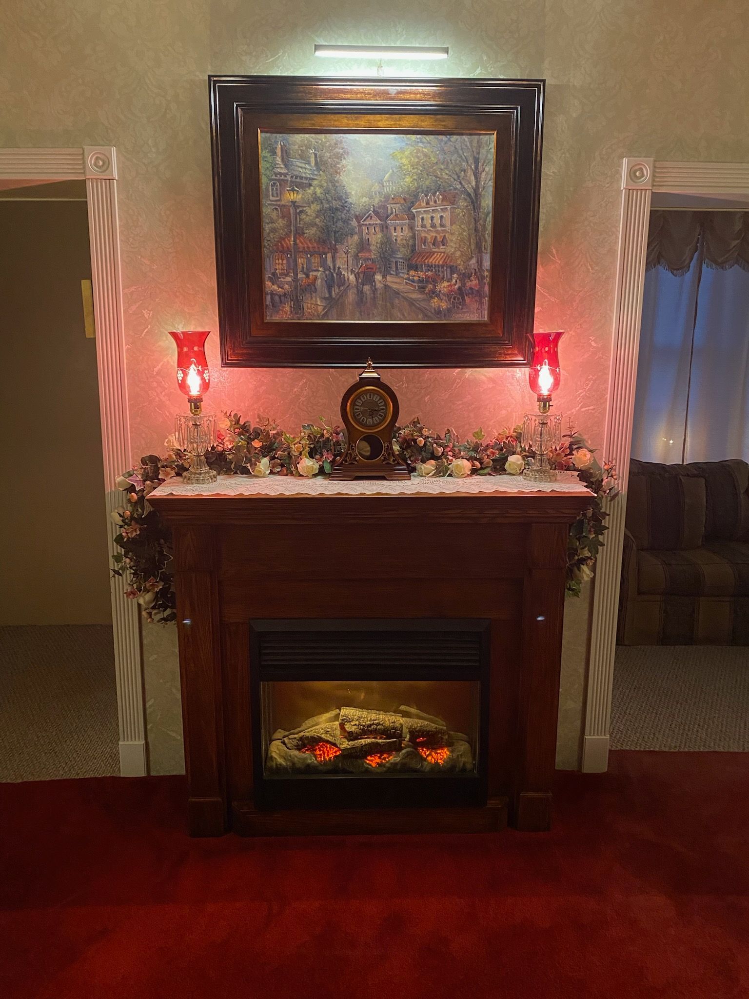 A fireplace mantel with a painting above it, flanked by two red lamps and decorative garland, in a room with red carpet.