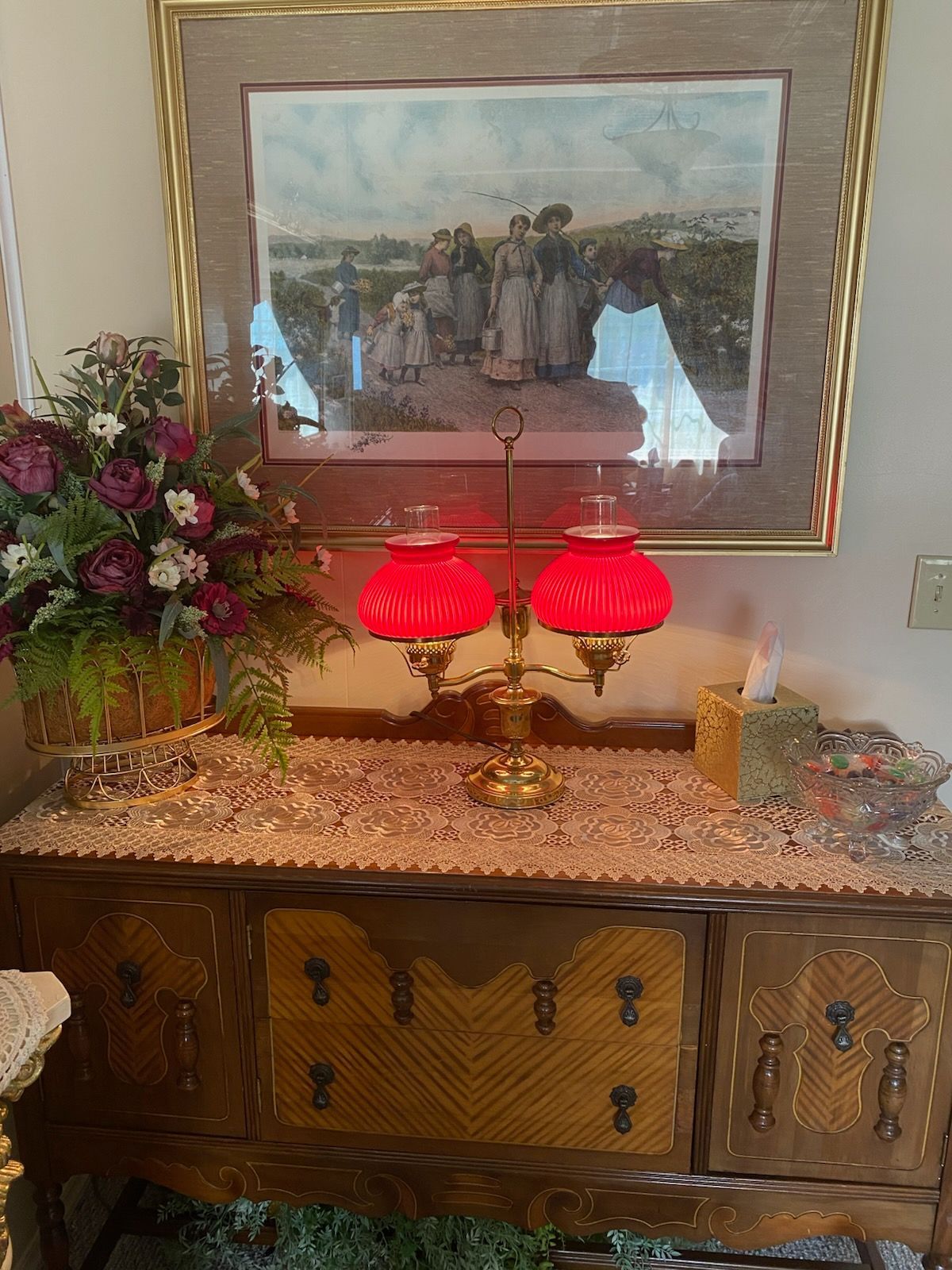 A vintage wooden sideboard topped with a dual red-shaded lamp, a floral arrangement, and a framed print on the wall above.