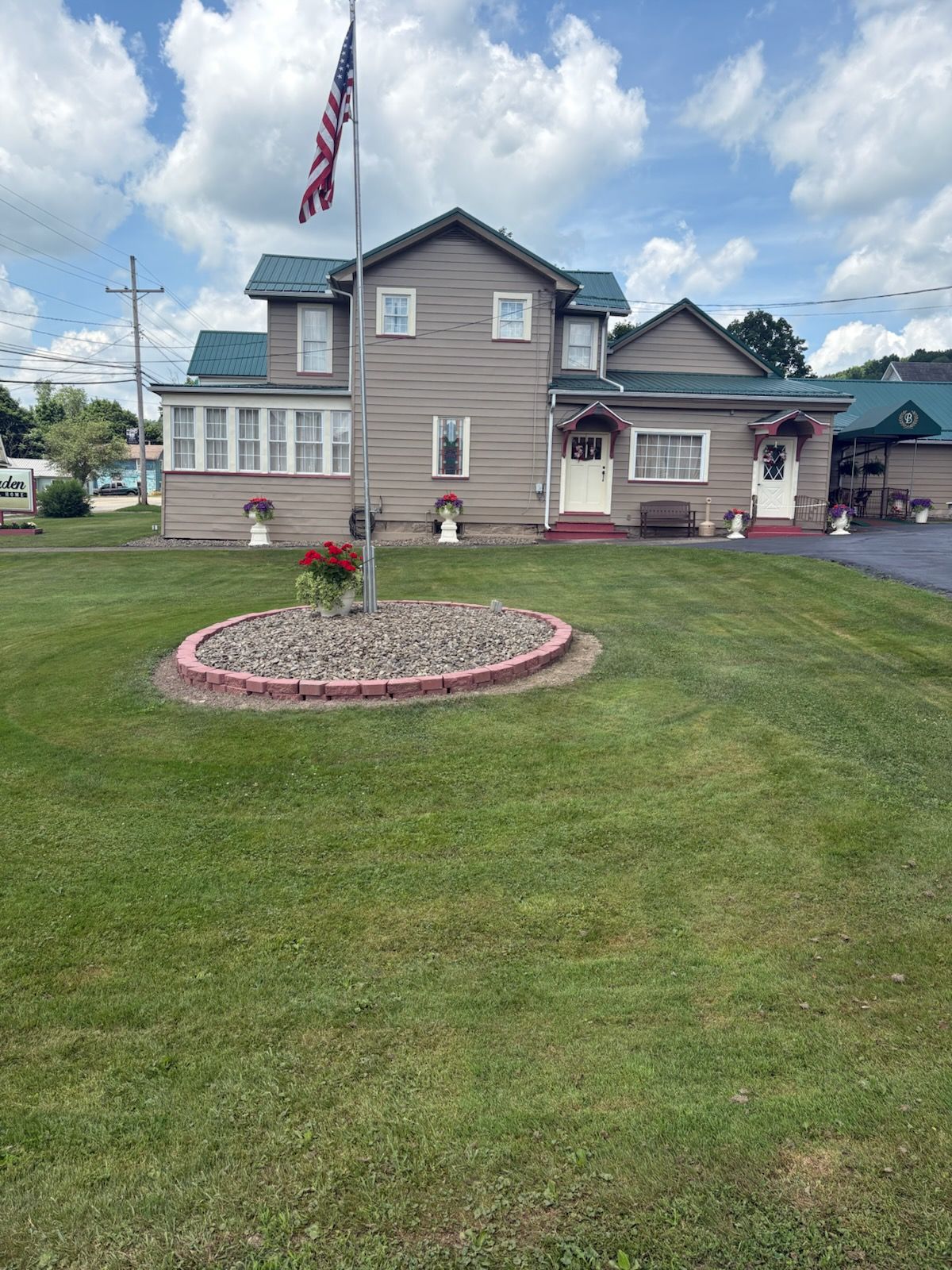 A tan two-story house with a green roof, American flag, and a circular stone flower bed on a green lawn.