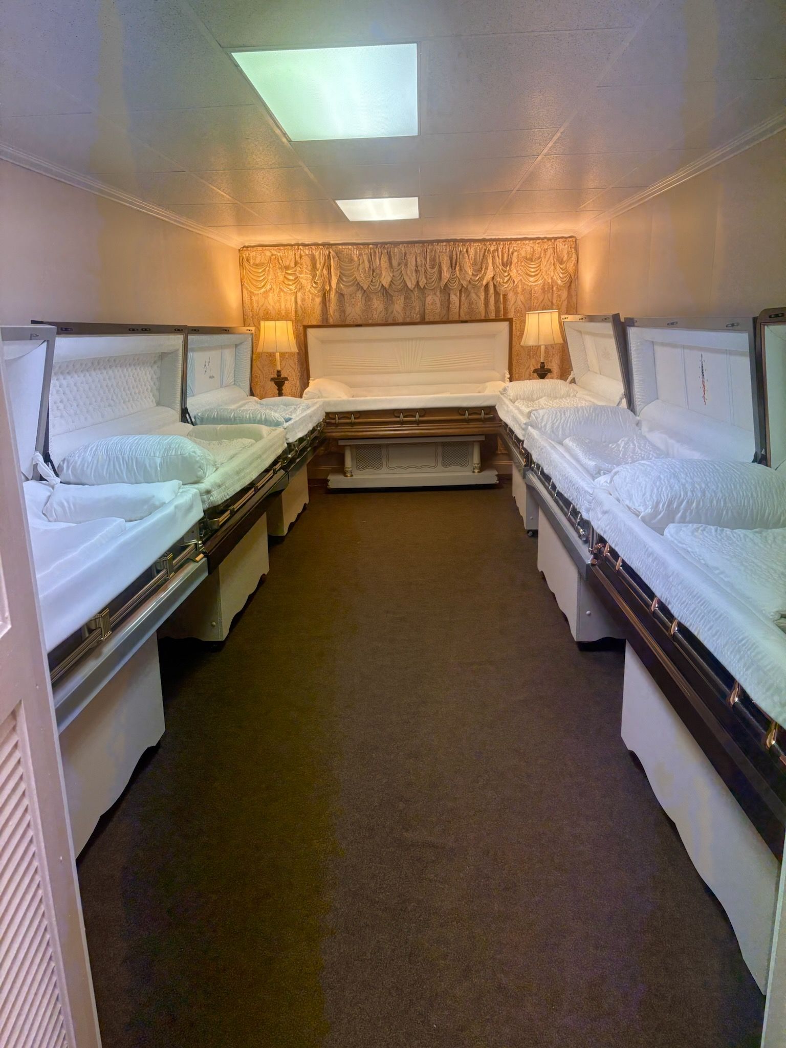 A funeral home display room features several open, white-lined caskets arranged on both sides of a carpeted aisle.